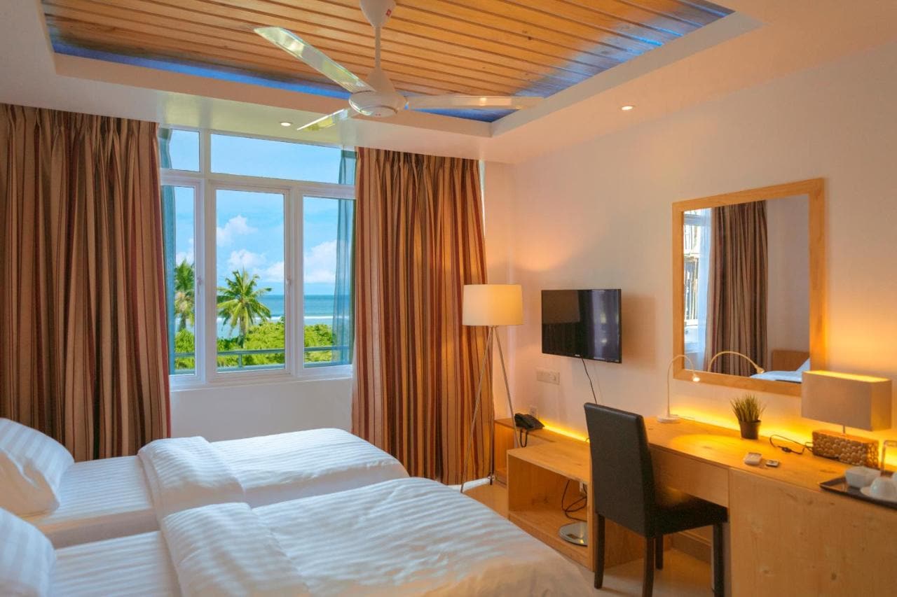 Deluxe Double room with white bedding, a desk and mirror, and a large window with palm trees outside